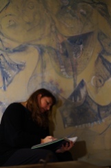 Maria L. Berg writing in her notebook in front of her portrait painted by E. Spencer Matthews entitled Maria Fights the Robot Spiders